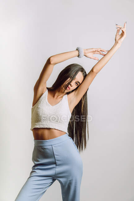 Trendy giovane donna etnica in crop top e braccialetto guardando la fotocamera mentre in piedi con braccio sollevato su sfondo chiaro — Foto stock