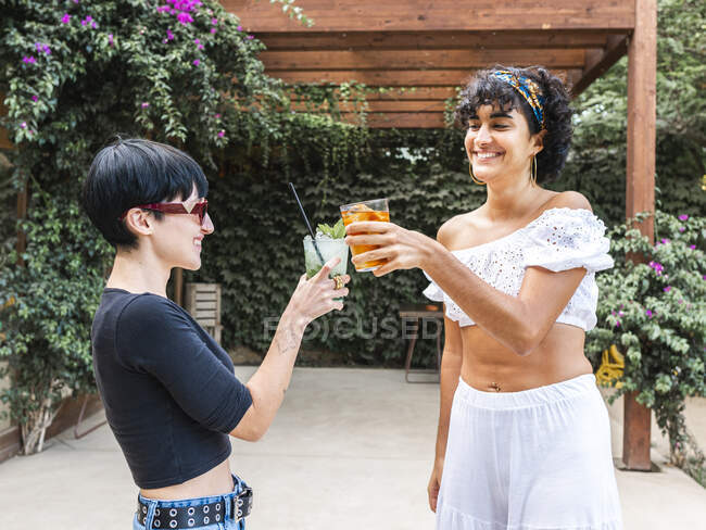 Веселые многонациональные подруги, звонящие стаканы холодных освежающих коктейлей, отдыхая вместе на террасе в летнем саду — стоковое фото