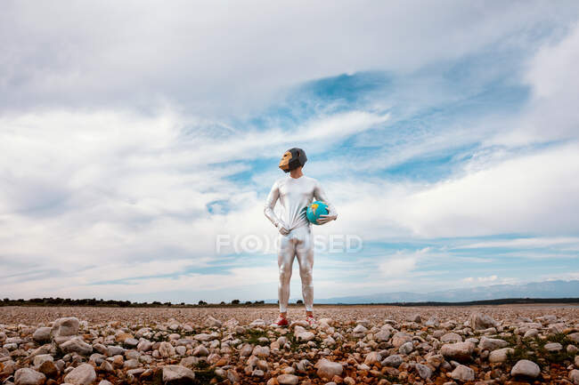 Анонимный парень в серебряном латексном костюме с геометрической обезьяньей маской, смотрящий в сторону и держащий глобус на природе — стоковое фото