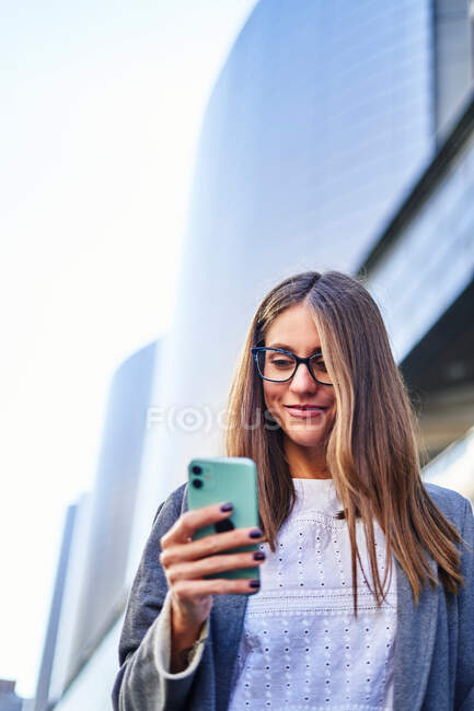 Знизу позитивна жінка в офіційному носінні використовує мобільний телефон під час прогулянки по міській вулиці для роботи — стокове фото