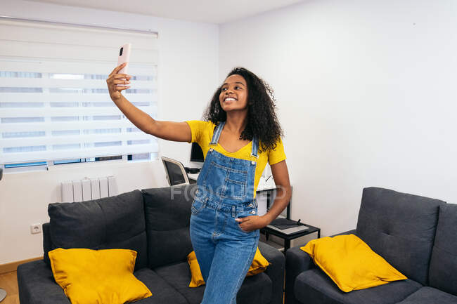 Счастливая афроамериканка блогер с длинными вьющимися волосами в желтой футболке и джинсах, стоящая с рукой в кармане и делающая селфи на смартфоне в светлой комнате — стоковое фото