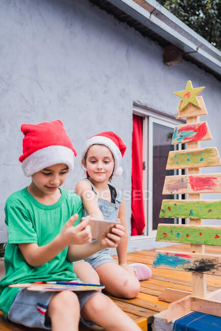 Enfants en chapeaux de Père Noël rouge naviguant sur le téléphone portable tout en étant assis près décoratif peint arbre de Noël dans la pièce lumineuse pendant la célébration des vacances — Photo de stock
