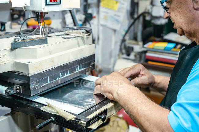 Vista lateral del atento maestro masculino senior de cultivo en delantal y gafas graduadas que operan la máquina laminadora en el taller de impresión - foto de stock