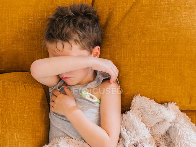 Зверху хворий хлопчик вимірює температуру за допомогою електронного термометра, лежачи на дивані вдома і маючи грип — стокове фото