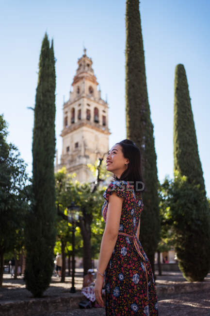 Боковой вид оптимистичной азиатской туристки, стоящей на улице с высокими зелеными деревьями и средневековым зданием в Испании в летний день — стоковое фото