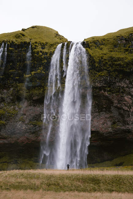 Vista posteriore di anonimo viaggiatore maschile in abito caldo in piedi e ammirare pittoresca rapida cascata Seljalandsfoss sotto cielo nuvoloso in Islanda — Foto stock