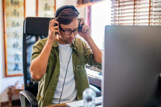 Чоловік одягає навушники, сидячи на стільці за столом і працюючи на комп'ютері біля вікна — стокове фото