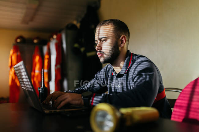 Concentrated young bearded ethnic guy wearing shirt sitting at table with laptop and transmitter looking at screen with yellow flashlight in foreground — Stock Photo