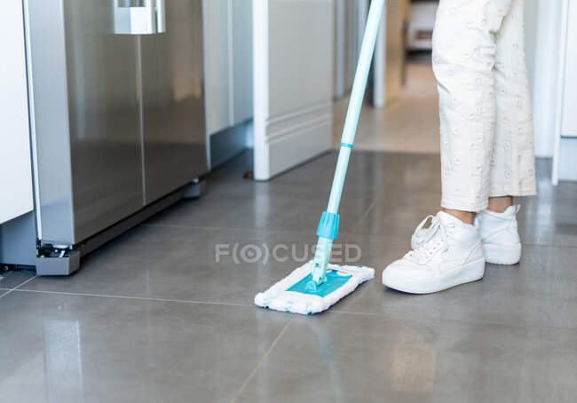 Vista lateral da safra senhora anônima em roupas casuais e tênis lavando piso em azulejo com esfregão de microfibra na cozinha leve — Fotografia de Stock
