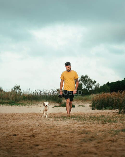Corps complet du propriétaire mâle avec chien obéissant se promenant sur le rivage sablonneux contre les plantes vertes le jour d'été dans la nature — Photo de stock