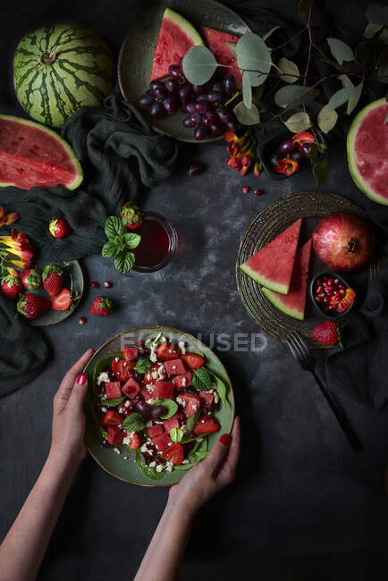 Draufsicht der Ernte anonyme weiblich mit Teller Wassermelonen-Salat mit Erdbeeren auf schwarzem Hintergrund mit Granatapfel und Trauben platziert — Stockfoto