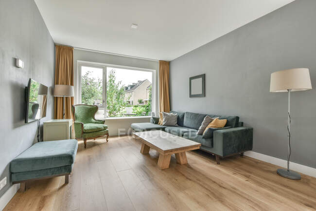Удобный диван рядом с деревянным столом, расположенным рядом с креслом у стены с телевизором в светлой гостиной с окном в квартире — стоковое фото
