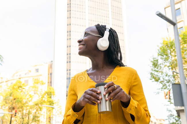Femme afro-américaine joyeuse avec thermo mug dans les mains écouter des chansons dans les écouteurs dans la rue avec le bâtiment et les arbres — Photo de stock