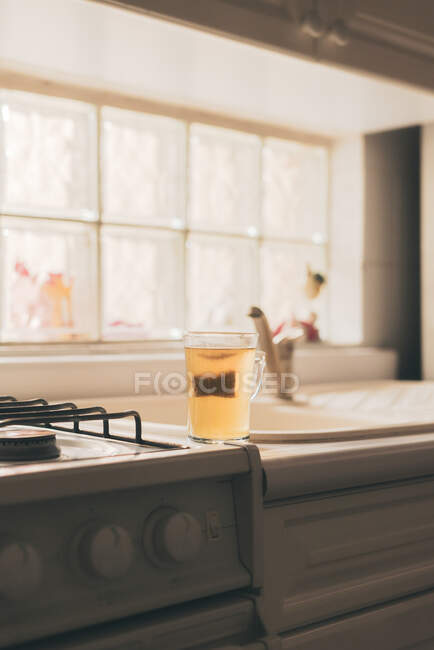Tazza di vetro con tè verde preparato in sacchetto posto sul bordo della stufa a gas in cucina — Foto stock