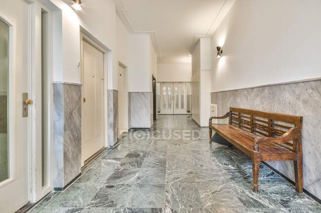 Aged shabby wooden bench placed in hallway of spacious building with marble tiled walls and white doors — Stock Photo
