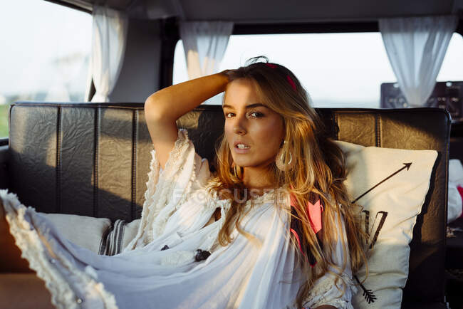 Atractiva chica rubia dentro de una furgoneta vintage y tumbada en el asiento en un día soleado mirando a la cámara - foto de stock