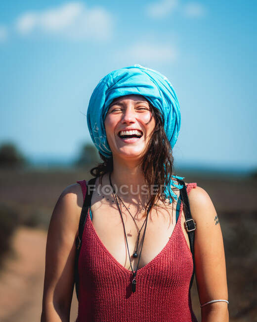 Позитивна жінка з коричневим волоссям в повсякденному одязі і хустці стоїть і сміється в сільській місцевості в сонячний день — стокове фото
