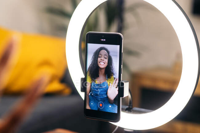Smiling black female on couch waving hand while using smartphone on LED ring lamp near professional lights on tripods — Stock Photo
