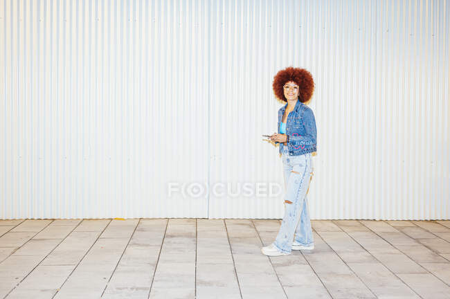 Full body of trendy female with Afro hairstyle wearing stylish outfit with sunglasses browsing cellphone while standing on street near wall — Stock Photo