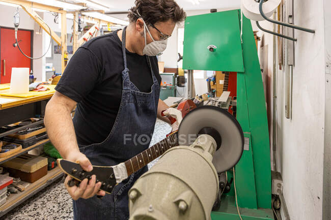 Вид сбоку серьезного мастера-мужчины в маске и рабочей одежде, шлифующего электрогитару на специальной машине во время работы в профессиональной мастерской — стоковое фото