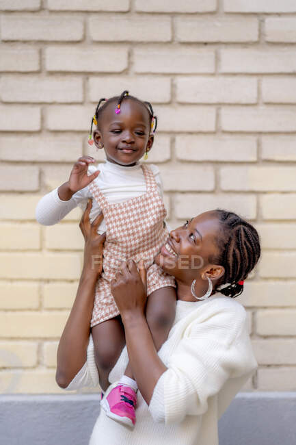 Fröhliche Afroamerikanerin mit Frisur steht und hebt kleine Tochter bei Tageslicht gegen Ziegelmauer auf der Straße — Stockfoto