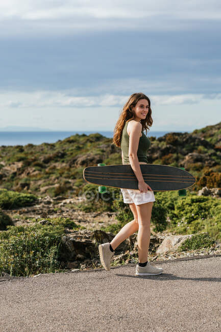 Side view of cheerful young female skateboarder with longboard strolling on walkway while looking away against mountain and sea — Stock Photo