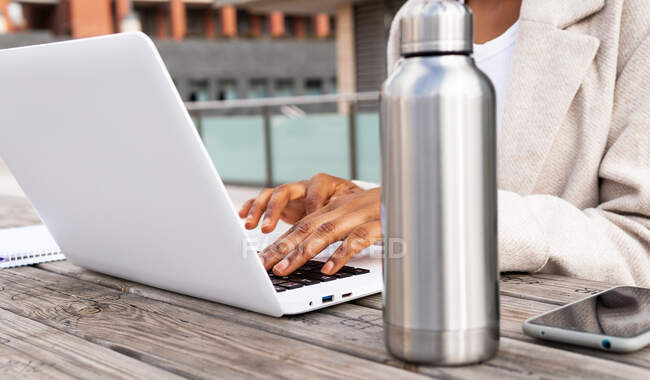 Side view of anonymous African American female freelancer browsing netbook while sitting on street at table with notebook during online work next to the thermos — Stock Photo