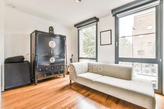 Comodo divano bianco posizionato vicino all'armadio nero invecchiato in ampio soggiorno con finestre che si affacciano su edifici residenziali in appartamento leggero — Foto stock