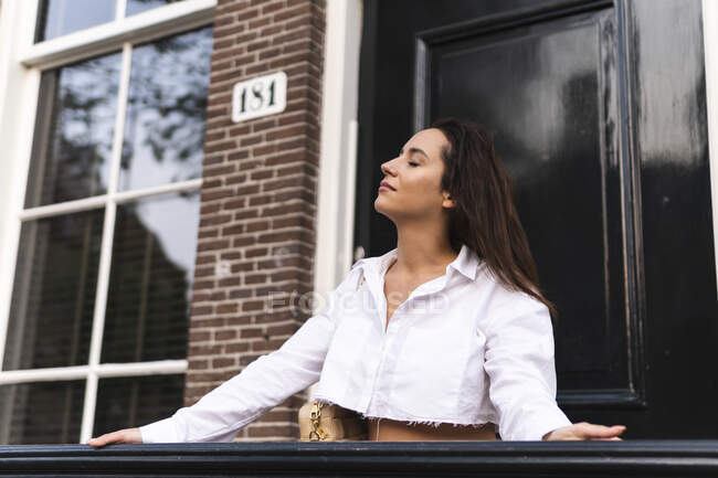 Mulher calma na roupa da moda em pé com os olhos fechados perto da entrada do edifício moderno com parede de tijolo e janela — Fotografia de Stock