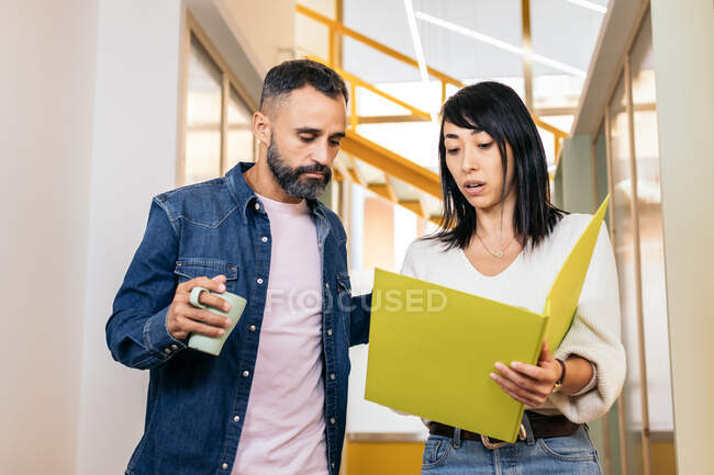 Empleada étnica concentrada en ropa casual mostrando una carpeta de documentos a un colega masculino serio parado en un espacio de coworking moderno con una taza de café - foto de stock