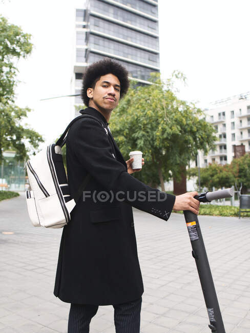 Giovane maschio afroamericano elegante con capelli ricci scuri in elegante cappotto in piedi sulla strada con scooter elettrico e tazza di caffè da asporto e guardando la fotocamera — Foto stock