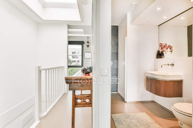 Corredor con valla y estante de madera cerca de baño elegante con lavabo blanco en el espejo y el inodoro en apartamento elegante luz - foto de stock