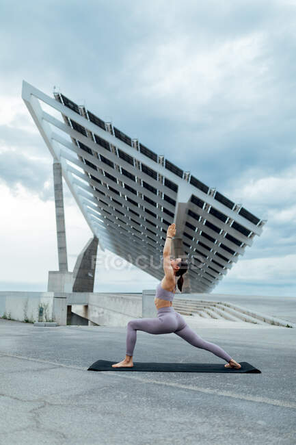 Full body of sportive female in activewear practicing Warrior posture while training on street near solar panel against cloudy sky — Stock Photo