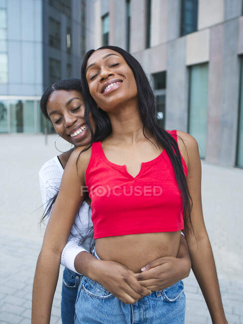 Positive lesbiche afroamericane in abbigliamento casual con gli occhi chiusi abbracciano mentre in piedi su strada con edifici moderni in città — Foto stock