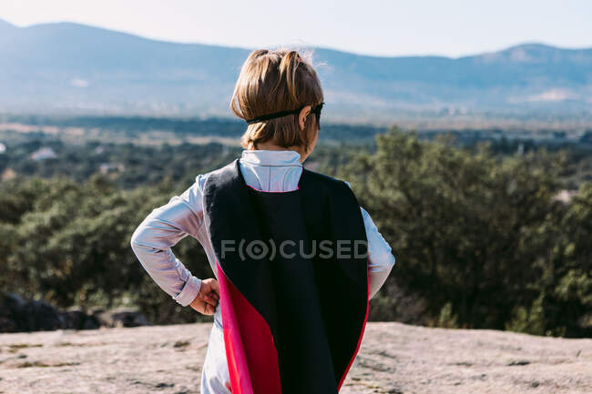 Visão traseira de pequena garota irreconhecível em traje de super-herói com as mãos na cintura em pé na colina rochosa — Fotografia de Stock