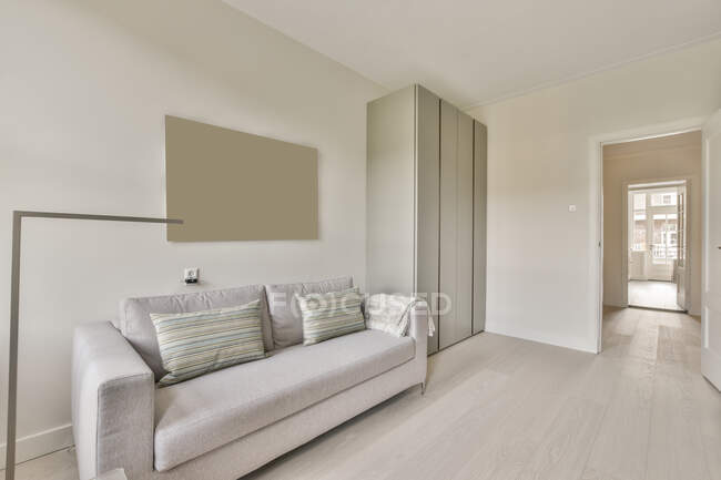 Cómodo sofá con cojines y armarios de estilo minimalista situado cerca de ventanas en la sala de estar soleada - foto de stock