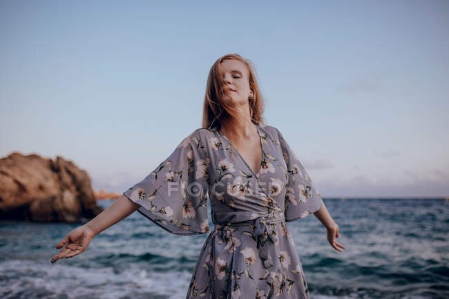 Mujer encantada con el pelo largo en vestido de moda de pie con los ojos cerrados en la orilla del mar en la noche de verano - foto de stock