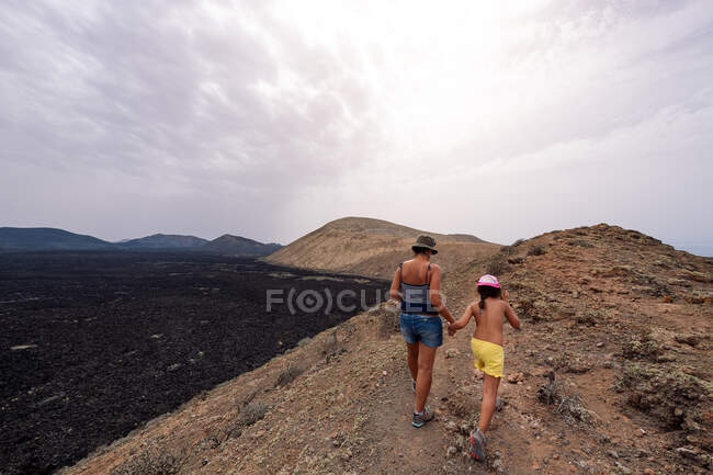 Rückenansicht einer anonymen Mutter mit einem Mädchen, das Händchen hält, während sie in malerischem Blick auf den Krater des Vulkans Caldereta gegen Berge und Meer unter hellem Himmel auf Lanzarote spaziert — Stockfoto