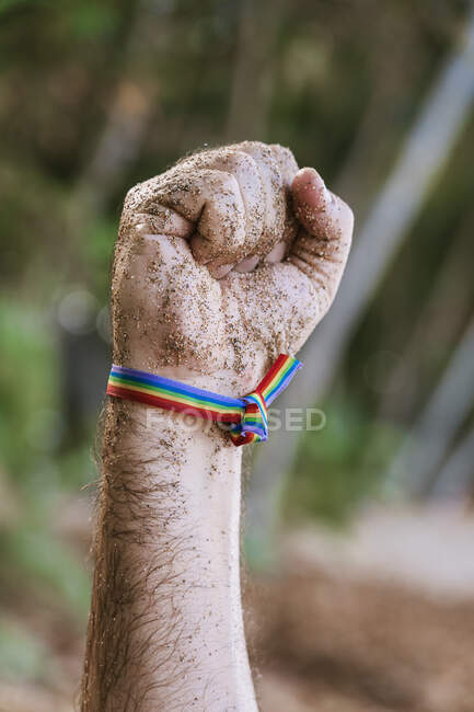 Врожай невизначений чоловік з веселковим браслетом, що демонструє кулак в піску проти зелених дерев, що ростуть в природі в сонячний день — стокове фото