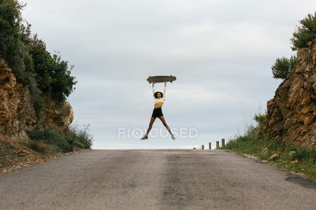Full body of positive ethnic female jumping high and raising hands with longboard on asphalt road — Stock Photo