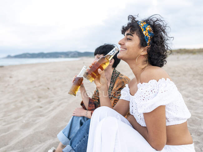 Позитивная многонациональная подруга, пьющая холодное пиво, сидя на песчаном берегу моря и смеясь летним вечером — стоковое фото