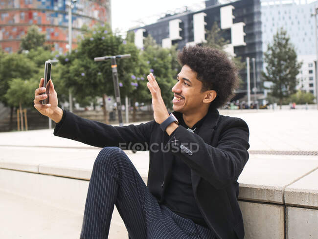 Веселий молодий чорний чоловік з темним афро волоссям в модному вбранні посміхається і махає рукою, маючи відеодзвінок через смартфон на міській площі — стокове фото