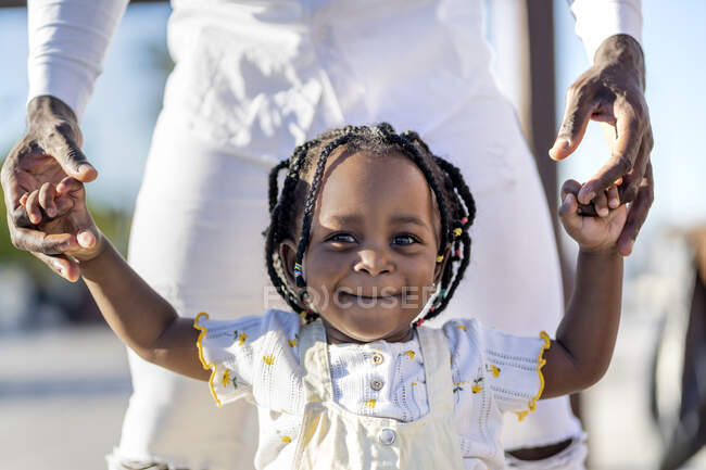 Feliz niña afroamericana en vestido ligero cogida de la mano de un padre irreconocible mientras camina por la calle en un día soleado - foto de stock