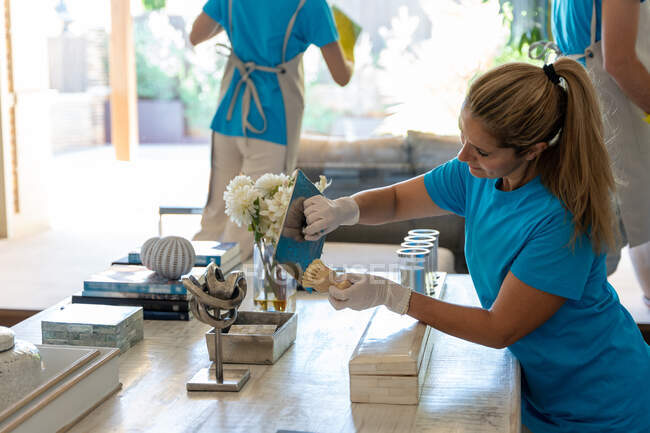 Side view of young female employee in uniform and gloves cleaning souvenirs on table with brush while working in spacious house with crop unrecognizable colleagues — Stock Photo