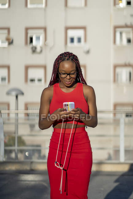 Cheerful African American female with braids in trendy red outfit standing and browsing smartphone on street against residential building in city in sunlight — Stock Photo
