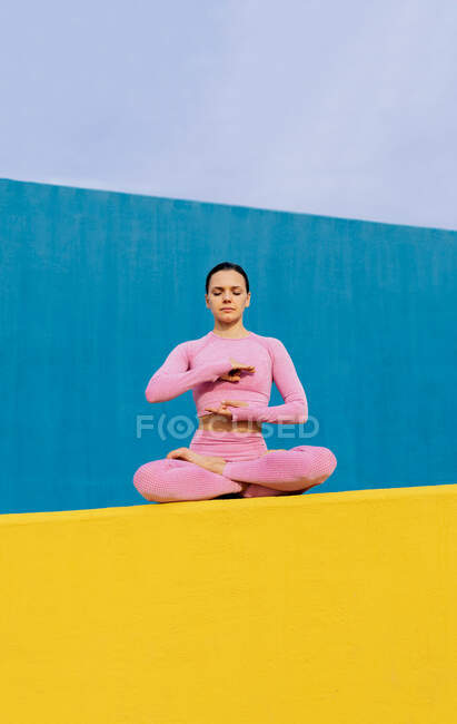 Спокойная гибкая женщина сидит в Падмасане и медитирует с закрытыми глазами, практикуя йогу на ярком синем и желтом фоне — стоковое фото