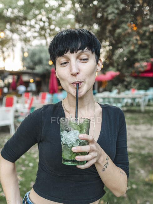 Содержание женщины, пьющей освежающий коктейль с кубиками льда и мятными листьями, стоя в летнем парке — стоковое фото