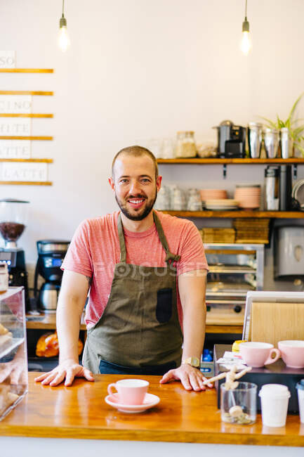 Barista masculin joyeux dans tablier souriant et regardant la caméra tout en se tenant au comptoir avec une tasse de café dans le café — Photo de stock