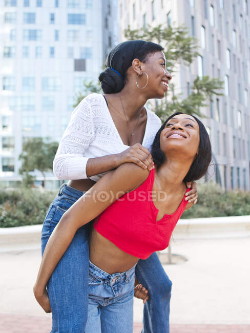 Delighted African American lesbian giving piggyback ride to content female partner while standing on street with modern buildings in city — Stock Photo
