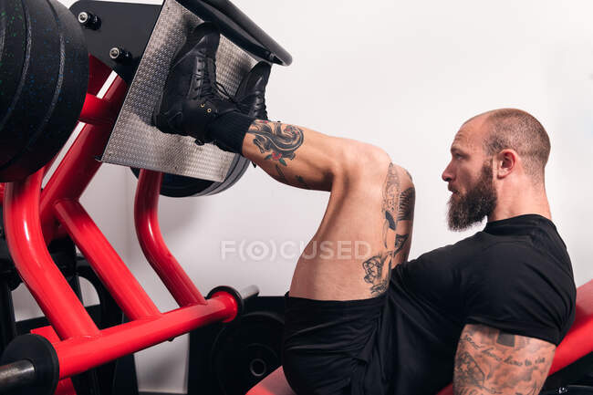 Seitenansicht eines muskulösen Sportlers mit Tätowierungen, der während des Trainings im Fitnessstudio Übungen an einem Beinpressgerät macht — Stockfoto